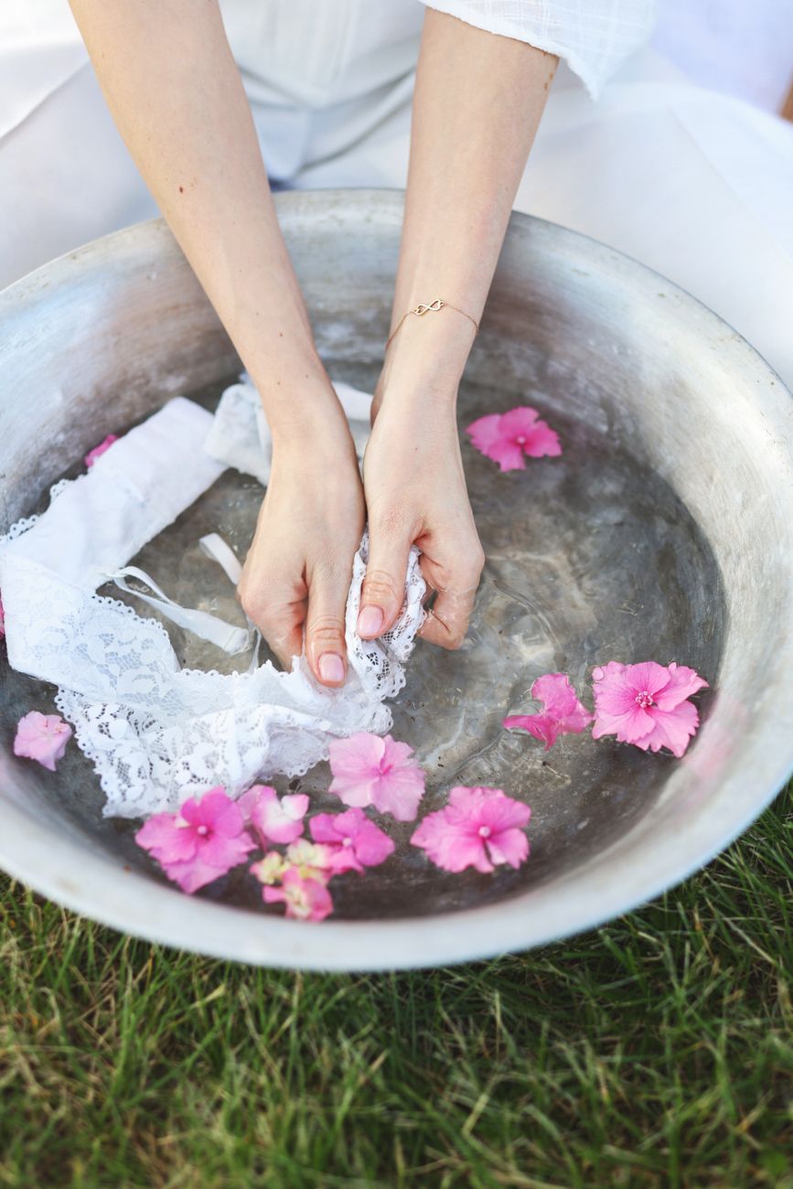 Close-up of hands washing white lace fabric in a metal bowl with pink flowers outdoors.