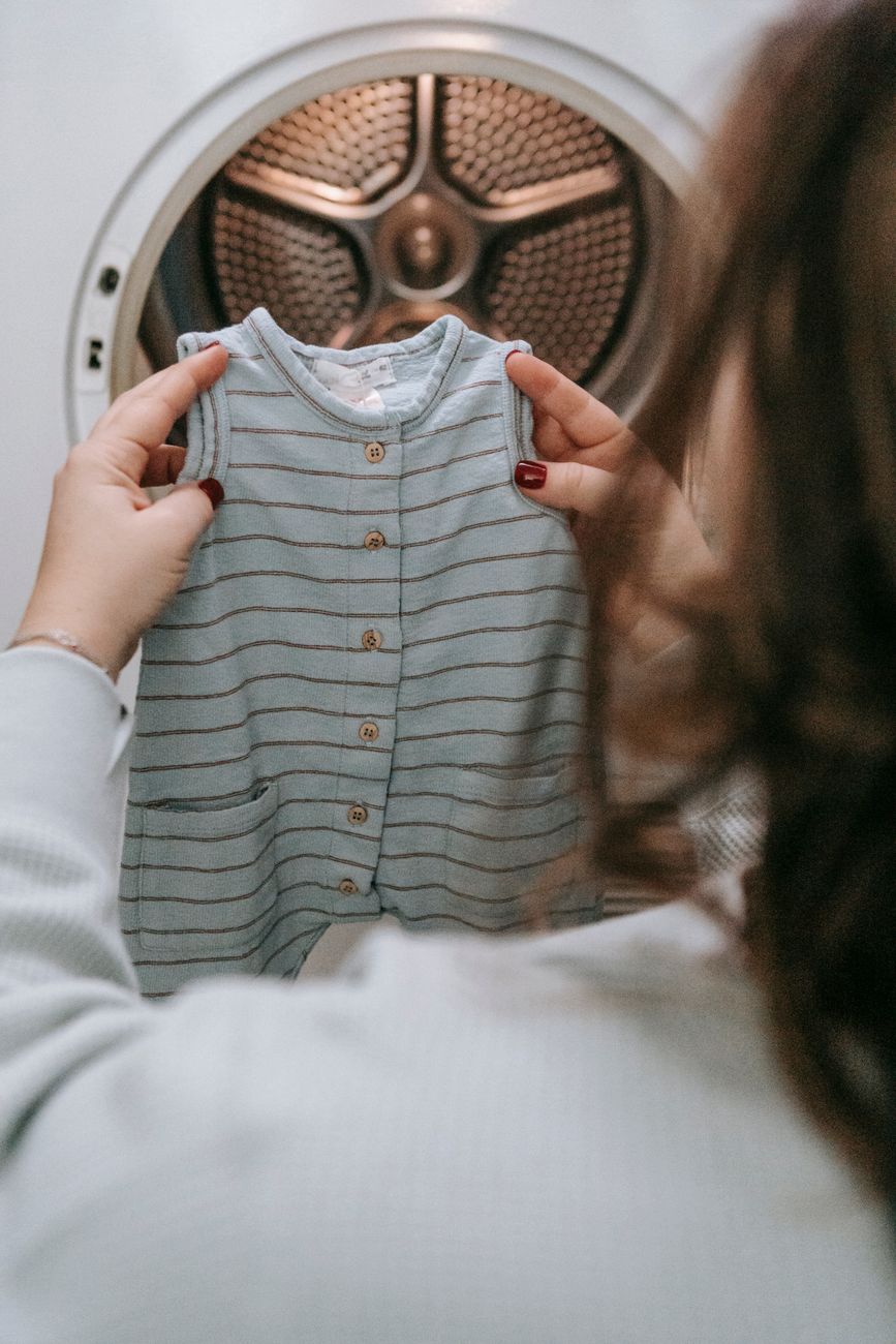 Back view of unrecognizable female with baby attire in hands near modern washing machine during laundry routine in light room